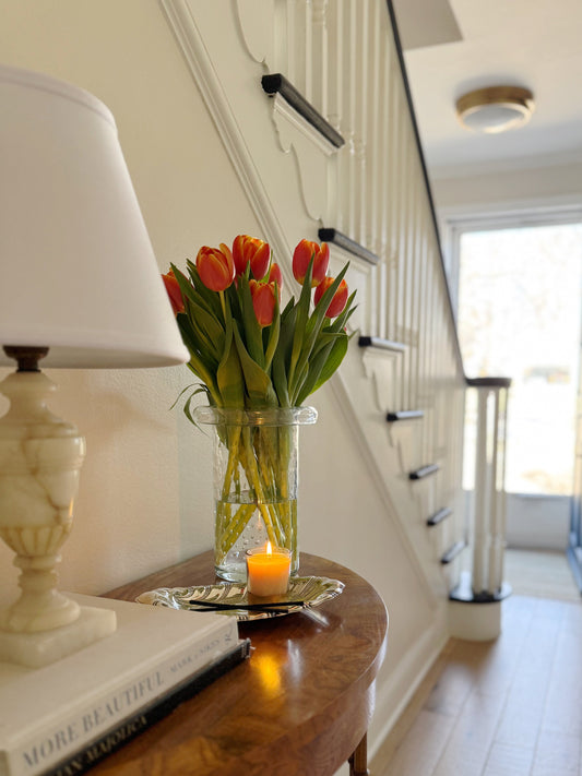 Vase of red tulips on a wooden table with a lamp and books in a room.